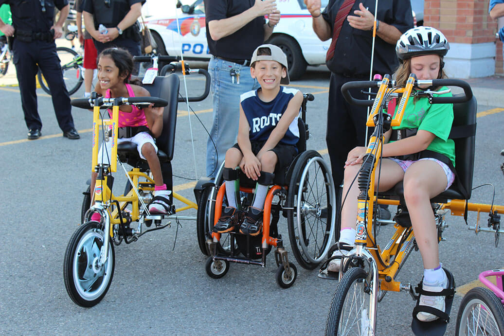Three children with disabilities in different pediatric wheelchairs having fun outside at an event.